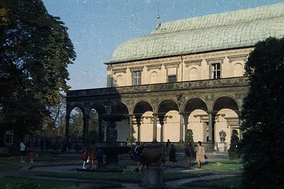 Ornate, 18th-century palace features a grand arcaded facade and copper roof, viewed from a manicured garden. Visitors stroll ...