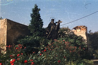Statue of a robed figure, possibly a monk or saint, stands amidst vibrant red roses and overgrown foliage. A crumbling stone ...