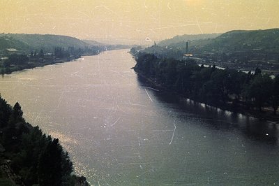 A wide river winds through a valley flanked by dense evergreen forests. A distant industrial structure and a few buildings ar...