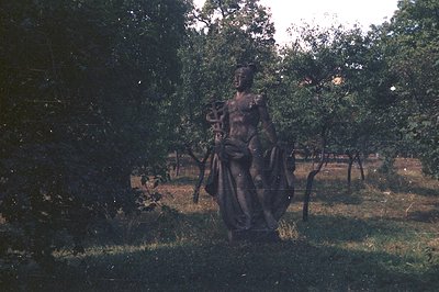 A bronze statue depicting a male figure, likely Asclepius, holds a staff entwined with a snake. Set within a grassy parkland,...