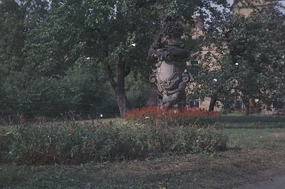 Ornate stone monument stands within a manicured garden, framed by mature trees. Detailed carvings depict figures & heraldic e...