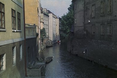 Narrow canal scene featuring tall, aged buildings lining both sides. Wooden boats are docked along the water’s edge. Appears ...