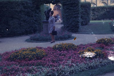 A woman in a light-colored coat and carrying a briefcase walks past manicured gardens and a bust statue. The vibrant floral d...