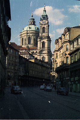 A view down a cobbled street showcases the baroque architecture of St. Nicholas Church in Prague. 1960s-70s cars line the roa...