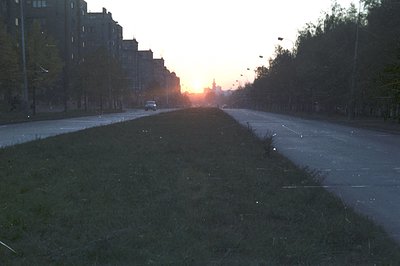 A wide avenue stretches towards a sunset, lined with tall, blocky apartment buildings. Lush green space fills the left side o...