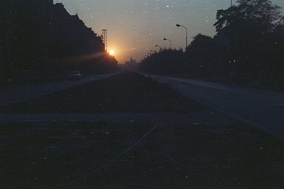 Long, straight street receding into the distance, with a single car visible. Sunset illuminates the sky, creating a hazy glow...