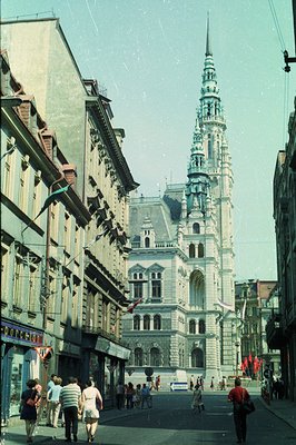A street view captures a grand, ornate town hall with a towering spire dominating the scene. Pedestrians populate the street ...
