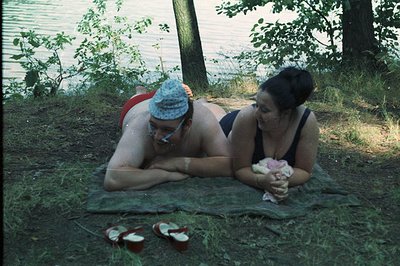 A couple rests on a blanket near a waterfront, possibly a lake or river. The man wears a patterned swimming cap & sunglasses;...