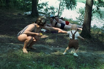 A barefoot woman in a dark striped swimsuit reaches for a toddler wearing overalls and sneakers. Two women in swimwear reclin...