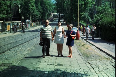 Three figures walk hand-in-hand along a cobbled street lined with trees and a tram line. An older man in striped shirt, a wom...