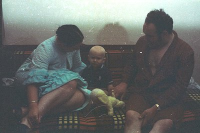A seated family—woman in a blue quilted robe, child in a black sweater, and man in a brown robe—captured in an interior space...