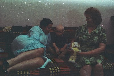 A young child with a bald head sits cradling a stuffed lamb, flanked by two women. The scene appears domestic, likely indoors...