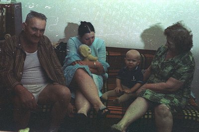 Family portrait: A man in striped pajamas, a woman in a blue robe, and a baby sit on a floral sofa. A teddy bear rests in the...
