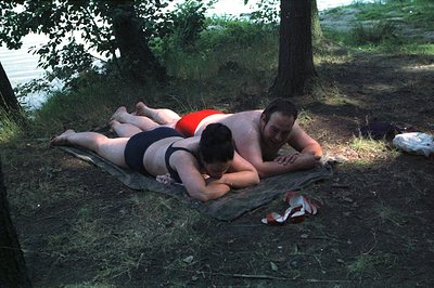 A couple lounges on a worn green blanket near a waterfront, possibly a lake or river. The man, in a red swimsuit, rests his h...