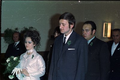 A newlywed couple stands amidst guests, the bride in a vintage, textured dress and bouquet. The groom wears a dark suit and t...