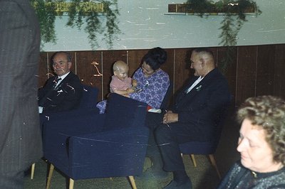 Formal portrait showing three adults and a baby seated in dark blue armchairs. The setting features wood paneling and hanging...