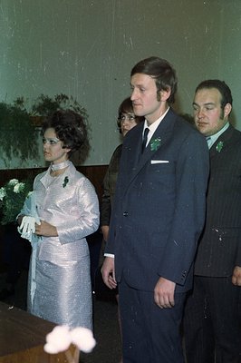 Three formally dressed individuals stand in a photograph, likely at a wedding or formal event. The woman wears a shimmering, ...