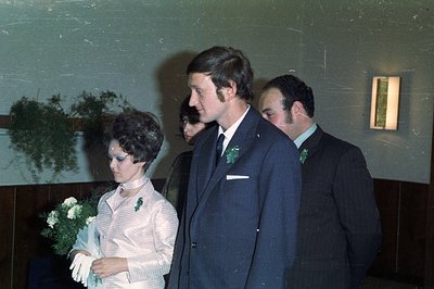 A candid color photograph, likely from the 1970s, depicts a wedding party. A bride, holding a bouquet of white flowers, walks...