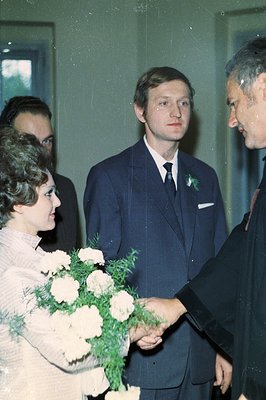A formal portrait from the 1970s depicting a young man in a dark suit & tie, mid-handshake. A woman in a matching sweater pre...