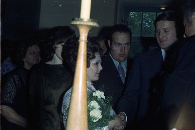 A young woman in a white dress & bouquet stands between two men in suits. Guests blurred in the background, suggesting a form...