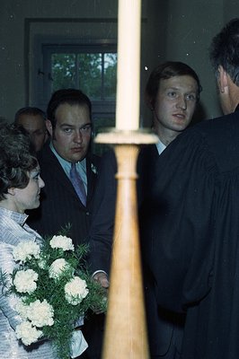 A formal indoor gathering; a young woman holds a bouquet of white carnations, while men in suits and a graduation robe stand ...