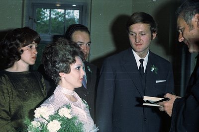 A posed group portrait likely captures a wedding event in the 1970s. The central figure holds a bouquet of white flowers, wea...