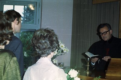 A young man in a suit and a woman in a green dress stand facing a clergyman during what appears to be a religious ceremony. T...