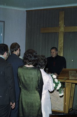 A solemn scene captures a church service. A figure in clerical robes presides, alongside two formally dressed men, and a woma...