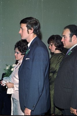 A formal portrait captures a wedding party, likely 1970s. Three individuals are seen in profile: a groom in a navy suit, a br...
