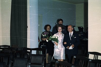 A bride in a midi-length dress with a floral bouquet walks with an older man, likely her father, down a church aisle. Two add...
