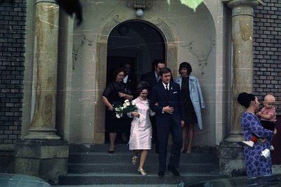 A couple exits a building, likely a wedding, framed by ornate columns and steps. The woman wears a short, pink dress & carrie...