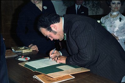 A man in a dark pinstripe suit signs a document on a green-covered table, visible in profile. Two women in formal attire stan...