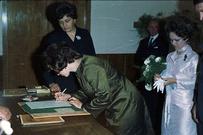 A woman in a dark green dress signs a document at a table, framed by formal guests. Note the 1970s fashion—shimmering dress, ...