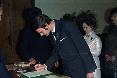 A man in a tuxedo signs a document; likely a wedding certificate. A woman with large, curled hair and a shimmering gown stand...