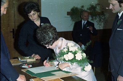 A woman signs a document, likely a marriage certificate, with a floral bouquet held in her arms. Two men in suits and a woman...