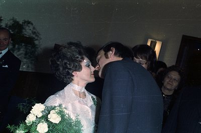 A couple shares a kiss, likely at a wedding reception. The bride wears a textured dress and carries a bouquet of white flower...