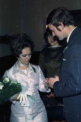 A bride in a pale blue suit dress holds a bouquet as a man, in a dark suit and tie, places a ring on her finger. A woman obse...