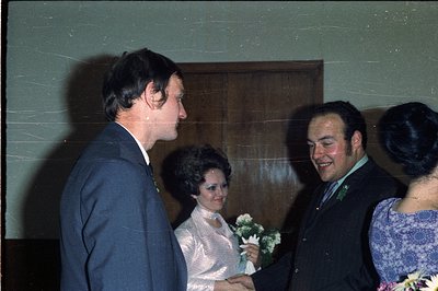 Three individuals in formal wear stand in a darkened interior, likely a hallway or entrance. A man in a dark suit speaks to a...