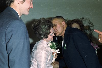 A candid moment at a wedding. A man in a dark suit leans in to kiss a woman in a patterned dress, likely the bride. A third p...