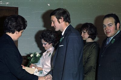 A woman presents a document to a formally attired couple. The bride wears a white dress, while the groom and supporting figur...