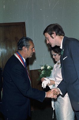A formally attired man presents a bouquet of flowers to a couple. The woman wears a shimmering dress and white gloves; the ma...
