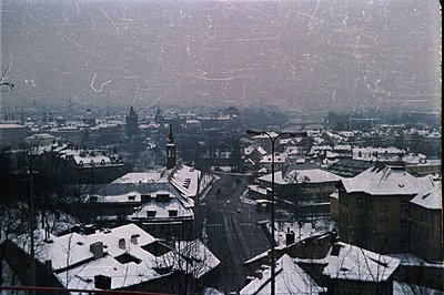 A panoramic view of a European city covered in snow, likely Central or Eastern Europe. Buildings display red-tiled roofs and ...