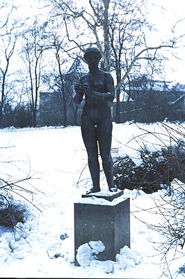 Bronze statue of a nude female figure stands atop a stone pedestal within a snow-covered garden. The figure holds an object i...