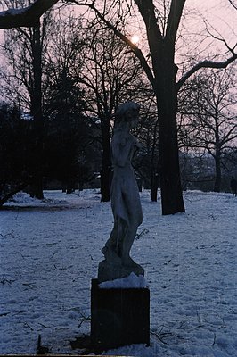 A snow-covered park scene featuring a classical sculpture of a draped woman atop a stone pedestal. Bare trees frame the view,...