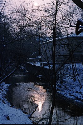 A partially frozen river reflects the setting sun in this wintry scene. Snow blankets the banks, contrasting with dark, bare ...
