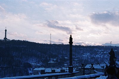 A snowy cityscape; rooftops capped with snow extend toward a wooded hillside. A tall, dark spire (likely a communications tow...