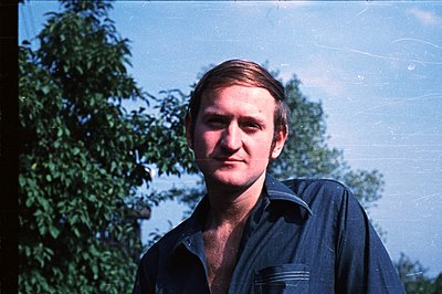 A portrait of a young man with short, neatly styled hair, wearing a dark blue work shirt. The background features dense folia...