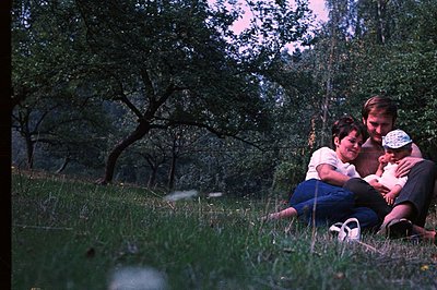 A young couple and baby sit in tall grass, surrounded by trees. The man, wearing a patterned shirt, holds the infant; the wom...