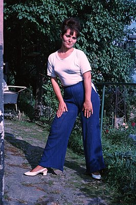 A young woman stands in a garden setting, wearing a white t-shirt and wide-legged, dark blue trousers. She’s captured in moti...