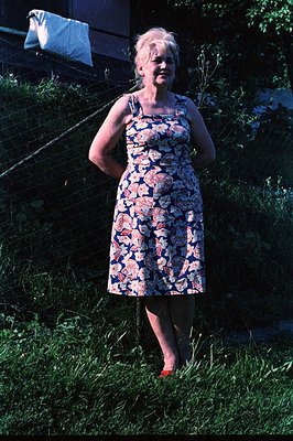 A woman in a floral print dress and red shoes stands on grassy ground. Clothes hang on a line behind her. Likely a domestic s...